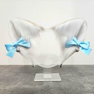 May include: White faux fur cat ears headband with light blue bows and silver bells. The headband is on a clear stand.