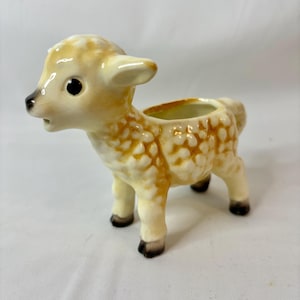 Può includere: Un vaso a forma di agnello in ceramica. L'agnello ha un corpo color crema con macchie marroni e occhi neri. Ha una piccola apertura sul dorso per piante o decorazioni.