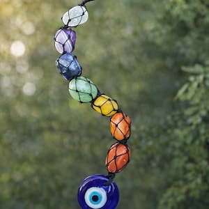 Może przedstawiać: Siedem zawieszek do tumble z czakrami z urokiem złego oka. Zawieszka zawiera szereg kolorowych, zaokrąglonych kamieni w siatce, z niebieskim złym okiem na dole. Tekst na zdjęciu mówi, że promuje równowagę, ochronę i uzdrawianie czakr.