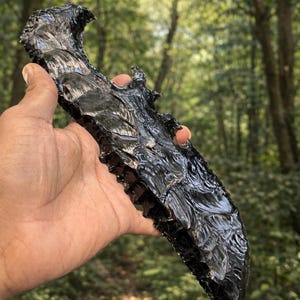 Könnte beinhalten: Eine handgehaltene, schwarze Obsidianklinge mit strukturierter Oberfläche und gezackter Kante. Die Klinge wird vor dem Hintergrund grüner Bäume gehalten, was ihre dunkle, reflektierende Qualität zeigt. Der Obsidian scheint handgefertigt zu sein.