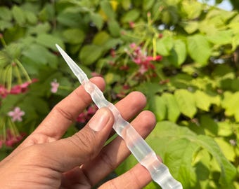 Könnte beinhalten: Ein klarer, länglicher Kristallstab mit einer Spitze und einem strukturierten, geriffelten Körper. Der Stab wird vor einem Hintergrund aus grünem Laub und rosa Blüten gehalten, was auf ein natürliches oder spirituelles Thema hindeutet.