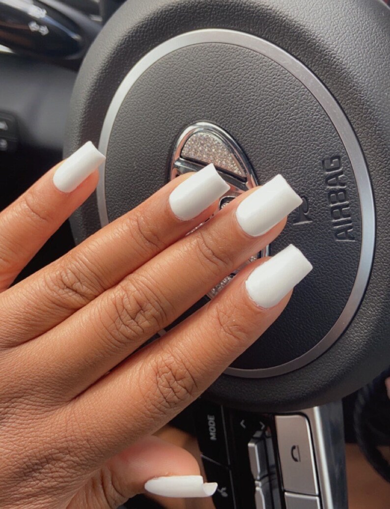 May include: Close-up of a hand with white square-shaped nails. The hand is resting on a black leather steering wheel with the word "Airbag" printed on it.