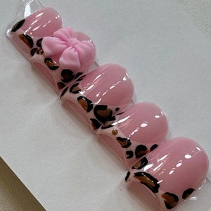 May include: Five pink press-on nails with a leopard print design. The nail on the left has a pink bow on top.