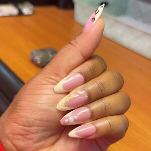 May include: Close-up of a hand with pink and white almond-shaped nails. The nails are decorated with gold leaf, pearls, and a white tip.