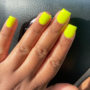 May include: Close-up of a hand with bright yellow square-shaped nails. The nails are painted a vibrant neon yellow.