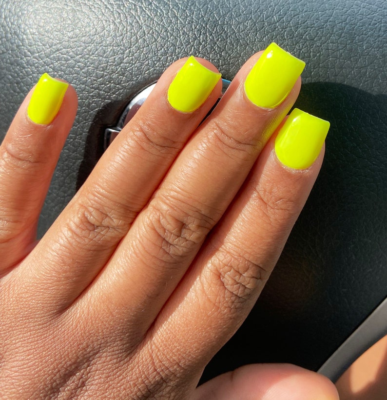 May include: Close-up of a hand with bright yellow square-shaped nails. The nails are painted a vibrant neon yellow.