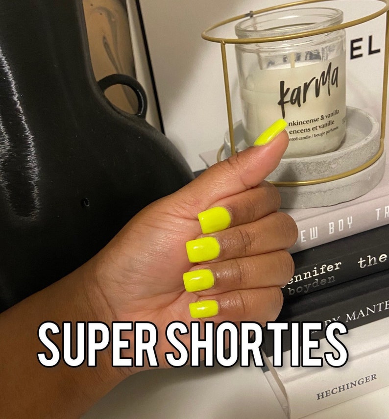 May include: Close-up of a hand with bright yellow short square-shaped nails. The hand is holding a thumb up. The background shows a candle with the word "karma" on the label and a stack of books with the words "New Boy" and "Super Shorties" visible.