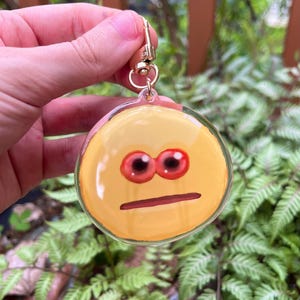 May include: A yellow round keychain with a surprised face with red eyes and a neutral mouth. The keychain is made of clear plastic and has a gold metal ring.