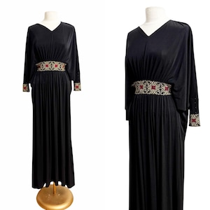 May include: A black maxi dress with a V-neck and long, flowing sleeves. The dress features an ornate, embroidered belt with red and gold accents. The cuffs also have matching embroidery. The dress is displayed on a mannequin.