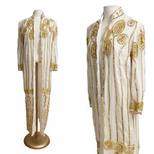 May include: A long white duster coat featuring vertical gold sequin stripes and intricate gold sequin swirl patterns along the collar, cuffs, and hem. The garment has a high neckline, long sleeves, and an open front design displayed on a mannequin.
