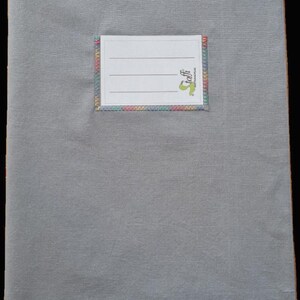 May include: A rectangular gray fabric book cover featuring a centered white label with three horizontal lines for writing. The label is stitched onto the gray fabric with a decorative, multi-colored zigzag thread border and includes the text toffi.