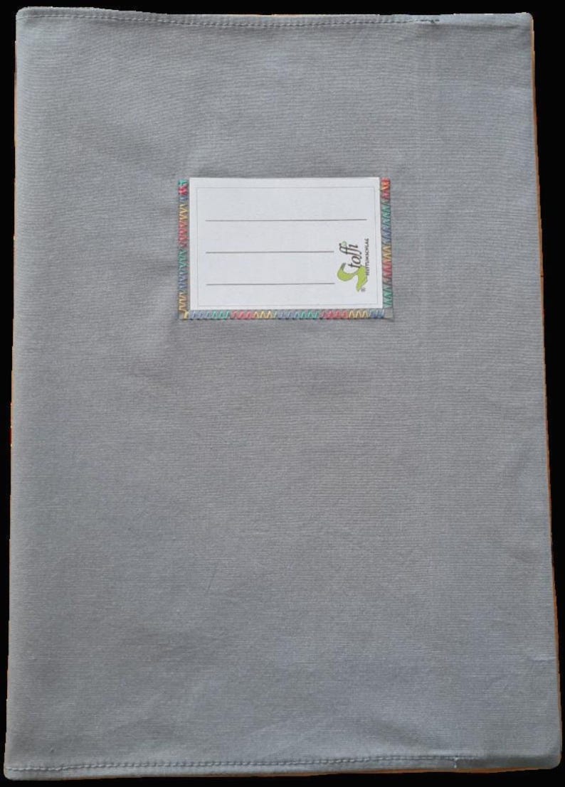 May include: A rectangular gray fabric book cover featuring a centered white label with three horizontal lines for writing. The label is stitched onto the gray fabric with a decorative, multi-colored zigzag thread border and includes the text toffi.