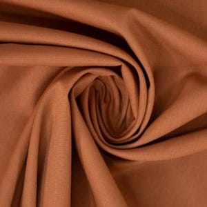 May include: A solid rust-colored fabric with a fine, woven texture. The material is gathered into a central spiral, creating deep, flowing folds and shadows that highlight the matte finish and consistent earthy tone of the textile surface.