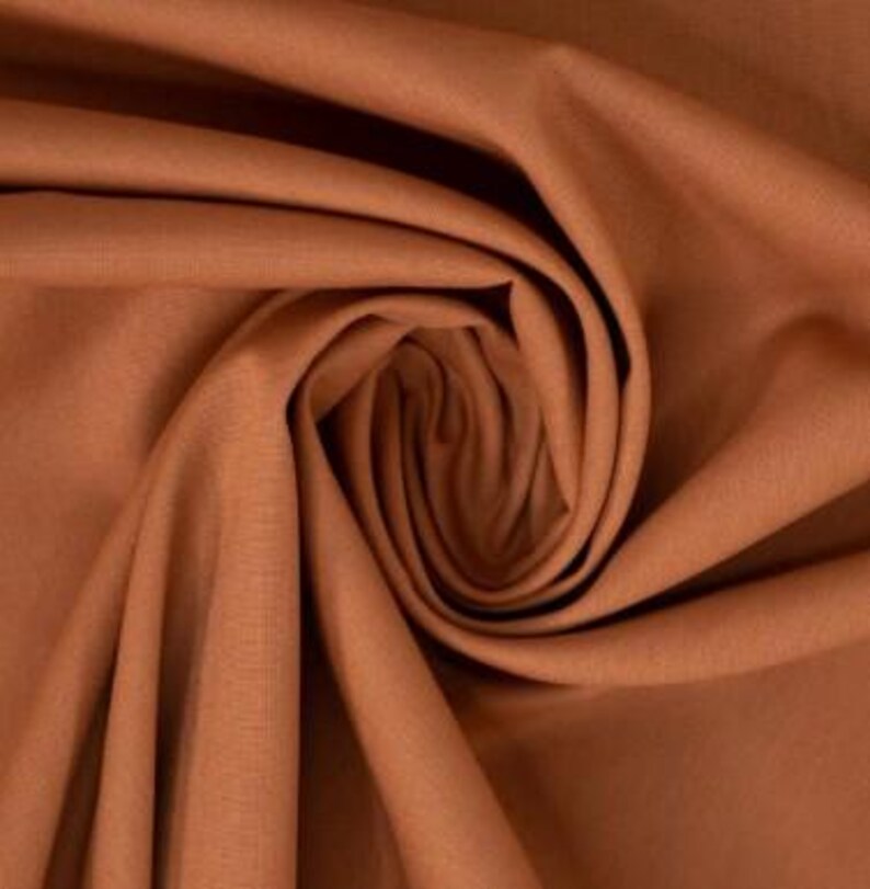 May include: A solid rust-colored fabric with a fine, woven texture. The material is gathered into a central spiral, creating deep, flowing folds and shadows that highlight the matte finish and consistent earthy tone of the textile surface.