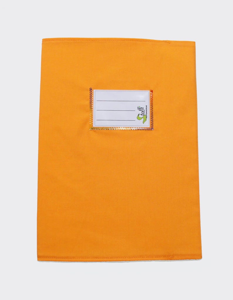 May include: A rectangular orange fabric book cover featuring a centered white label with three horizontal black lines for writing. The label is bordered by a metallic gold trim and includes the green text logo Stoffi printed on the bottom right corner.