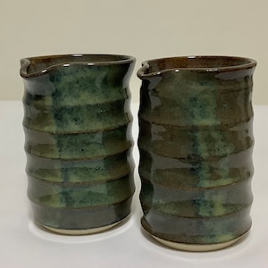 Op de afbeelding: Twee handgemaakte keramische kannen met een donkergroen en bruin glazuur. De kannen hebben een geribbeld ontwerp en een kleine tuit. De bodems zijn lichtbeige van kleur. De kannen worden waarschijnlijk gebruikt voor het serveren van dranken.