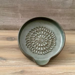 May include: A teal ceramic spoon rest with a floral pattern. The spoon rest has a circular design with a spiral centre and a small lip for holding a spoon. It is displayed on a wooden surface.