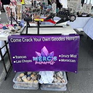 Puede incluir: Una pancarta morada y blanca con el texto "Come Crack Your Own Geodes Here!!" y el logotipo "MERC'S minerals". La pancarta está colgada de una mesa con dos contenedores de plástico transparente llenos de rocas de geodas marrones.
