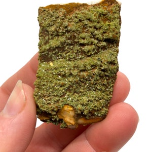 Puede incluir: Un primer plano de una muestra mineral verde y marrón con una superficie rugosa y texturizada. El mineral tiene un aspecto estratificado y está cubierto de pequeños cristales verdes.