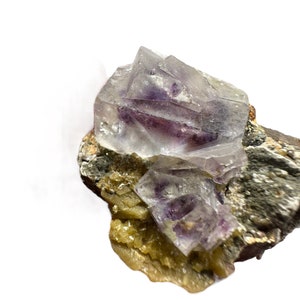 Peut inclure: Un groupe de cristaux de fluorine violets sur une matrice de roche grise et jaune. Les cristaux de fluorine sont clairs et ont une teinte violette.