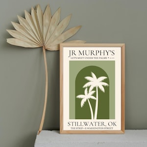 Op de afbeelding: Een ingelijste kunstdruk met een licht houten frame. De print toont een groene boog met twee witte palmbomen en de tekst "JR Murphy's Let's Meet Under The Palms" en "Stillwater, OK". Links is een gedroogd palmblad.