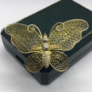 Może przedstawiać: Broszka w kształcie filigranowego motyla w kolorze złotym i srebrnym, prezentowana w zielonym pudełku z aksamitu.
