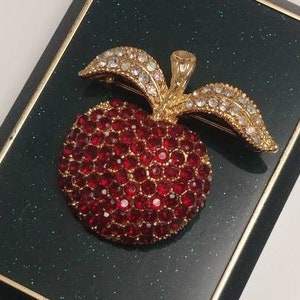 Puede incluir: Un broche de manzana roja con joyas, con detalles dorados y un tallo de cristal transparente. El broche se muestra sobre un fondo negro y verde.