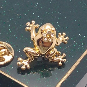 Może przedstawiać: Złota broszka w kształcie żaby z przezroczystymi cyrkoniami jako oczy. Żaba siedzi na zielonym tle.