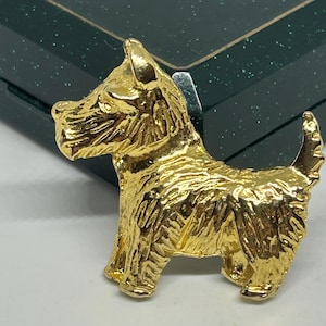 Może przedstawiać: Złota metalowa broszka w kształcie psa rasy Scottish Terrier. Broszka ma teksturowaną powierzchnię i stoi na czterech łapach. Ogon psa jest uniesiony, a broszka jest umieszczona na ciemnozielonym tle.