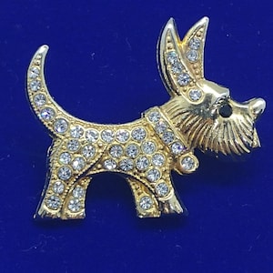 Może przedstawiać: Złota broszka w kształcie psa rasy terrier, wysadzana przezroczystymi cyrkoniami.