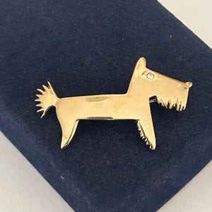 May include: A gold-toned metal brooch shaped like a Scottish Terrier dog. The brooch features a smooth, polished finish with a single small clear rhinestone set as the eye. The dog is depicted in profile with a pointed ear, a short tail, and a beard.