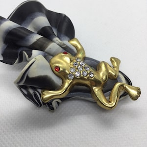 Może przedstawiać: Złota broszka w kształcie żaby z czerwonymi oczami i przezroczystymi cyrkoniami, umieszczona na czarno-białym materiale w kształcie spirali.