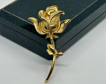 Könnte beinhalten: Eine goldfarbene Rosenbrosche mit einem detaillierten Design. Die Rose hat einen Stiel und Blätter.