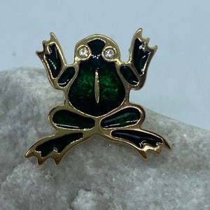 Może przedstawiać: Złota broszka w kształcie żaby z zieloną emalią i dwoma przezroczystymi cyrkoniami jako oczy.