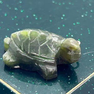 Könnte beinhalten: Eine kleine, grüne Jade-Schildkrötenfigur mit einer detailliert geschnitzten Schale und einem Kopf. Die Schildkröte sitzt auf einer grünen, glitzernden Oberfläche.