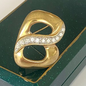 May include: A gold-toned brooch in the shape of the letter "B", embellished with a row of clear, sparkling rhinestones. The brooch is set against a dark green, textured background, suggesting a presentation box.
