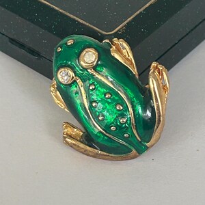 May include: A green frog-shaped brooch with gold-coloured accents. The frog has two clear, round rhinestones for eyes and small, gold-coloured dots on its back. The brooch is set against a dark green background.