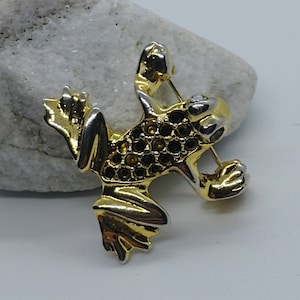 Może przedstawiać: Złota i srebrna broszka w kształcie żaby z czarnymi cyrkoniami. Żaba jest w pozycji skoku z wyciągniętymi nogami.