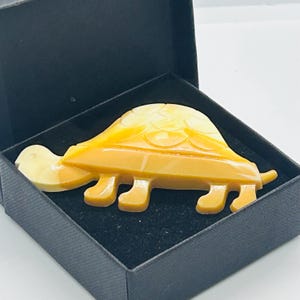 Może przedstawiać: Żółty broszka w kształcie żółwia z detalicznym wzorem skorupy. Broszka jest prezentowana w czarnym pudełku z aksamitu.