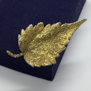May include: A gold leaf brooch with a textured surface. The leaf is detailed with small raised dots.