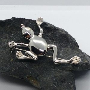 May include: A silver frog brooch with a shiny finish. The frog is in a leaping pose with its legs outstretched.