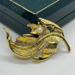 May include: A gold-toned leaf-shaped brooch with a single white pearl accent. The brooch is detailed with a textured surface and a delicate design.
