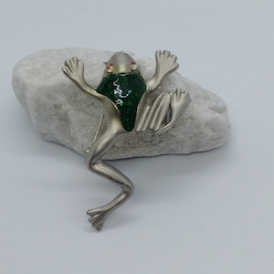 Może przedstawiać: Srebrna broszka w kształcie żaby z zielonym szkliwem i czerwonymi oczami. Żaba jest w pozycji z wyciągniętymi nogami i głową przechyloną na bok.