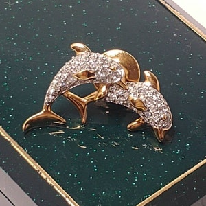 Może przedstawiać: Dwie złote figurki delfinów z przezroczystymi kryształowymi akcentami. Delfiny są zwrócone do siebie i umieszczone na zielonym tle.