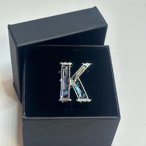 Op de afbeelding: Zilverkleurige letter "K" broche met iriserende abalone schelpinleg, gepresenteerd in een open zwarte geschenkdoos. De broche heeft een decoratieve rand en een gepolijste afwerking, geschikt voor persoonlijke versiering.