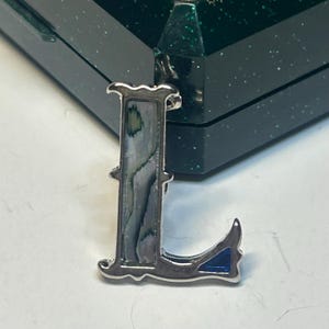 Op de afbeelding: Zilverkleurige letter "L" hanger met een abalone schelpinlegsel. De hanger heeft een decoratieve rand en een klein blauw emaille accent. De achtergrond bevat een donkergroene doos.