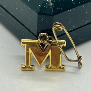 May include: Gold-toned letter M charm with a safety pin attached. The charm is on a green glittery surface.