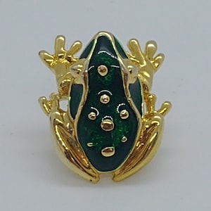 Może przedstawiać: Złota broszka w kształcie żaby z zielonym szkliwem. Żaba ma szczegółowy wzór z czarnymi oczami i fakturowanym ciałem.
