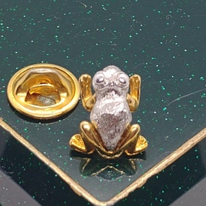 Może przedstawiać: Srebrna broszka w kształcie żaby z złotymi akcentami. Żaba siedzi na zielonej powierzchni z złotym brokatem. Broszka ma złote plecy z szpilką.