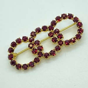 Może przedstawiać: Złota broszka z dwoma połączonymi owalnymi kształtami, każdy obramowany małymi, okrągłymi, ciemnoczerwonymi kamieniami szlachetnymi. Złota szpilka zabezpiecza broszkę. Projekt jest symetryczny i elegancki, odpowiedni na formalne okazje.